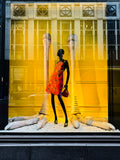 Black mannequin in a fashion storefront against a yellow backdrop, red floral dress, strong color contrast, urban fashion photograph with social commentary
